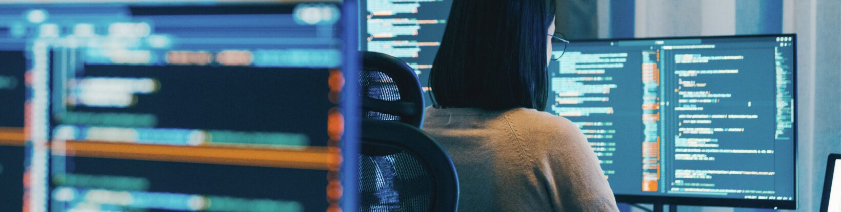 Person seated at a desk working on a computer with multiple monitors displaying lines of code.