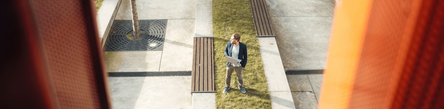 Person standing outdoors on a landscaped office campus, holding a laptop near benches and walkways.