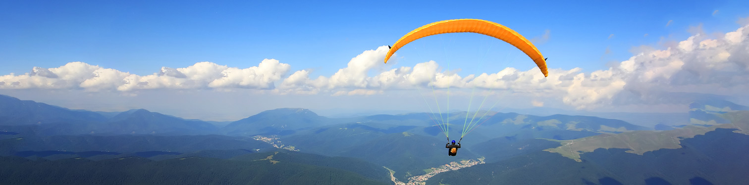 A single paraglider with an orange canopy flies high above a wide mountain range. Layers of blue and green hills stretch into the distance, with a river or road winding through a valley below. The sky is clear with scattered white clouds, and the paraglider is centered in the upper right portion of the image.