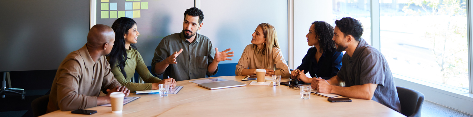 A diverse team of six professionals seated around a conference table, engaged in discussion during a collaborative meeting in a modern office.