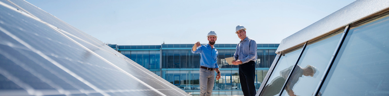 Two individuals in hard hats standing between rows of rooftop solar panels, with one pointing toward the panels while the other holds a tablet, set against a modern glass building.