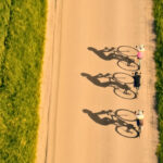 Aerial view of three cyclists riding in a line on a straight, light-brown paved road bordered by green grass on both sides, with long shadows cast to the left.
