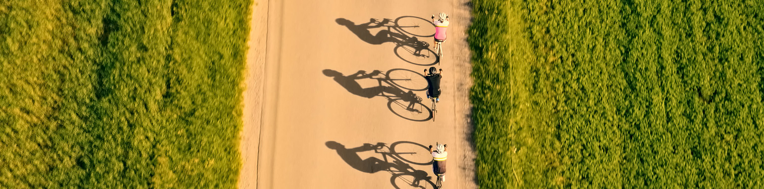 Aerial view of three cyclists riding in a line on a straight, light-brown paved road bordered by green grass on both sides, with long shadows cast to the left.