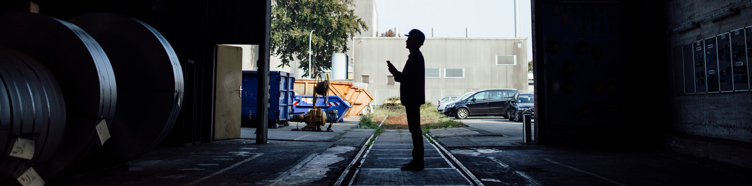 Silhouette of a worker wearing a hard hat standing inside an industrial facility, looking at a tablet near rail tracks and large equipment.