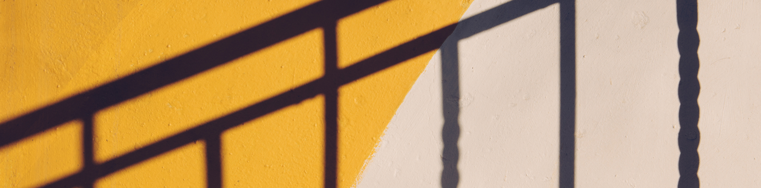 Dark shadows from a metal railing stretch diagonally across a textured wall painted in yellow and off-white, creating bold geometric lines and contrast in natural sunlight.