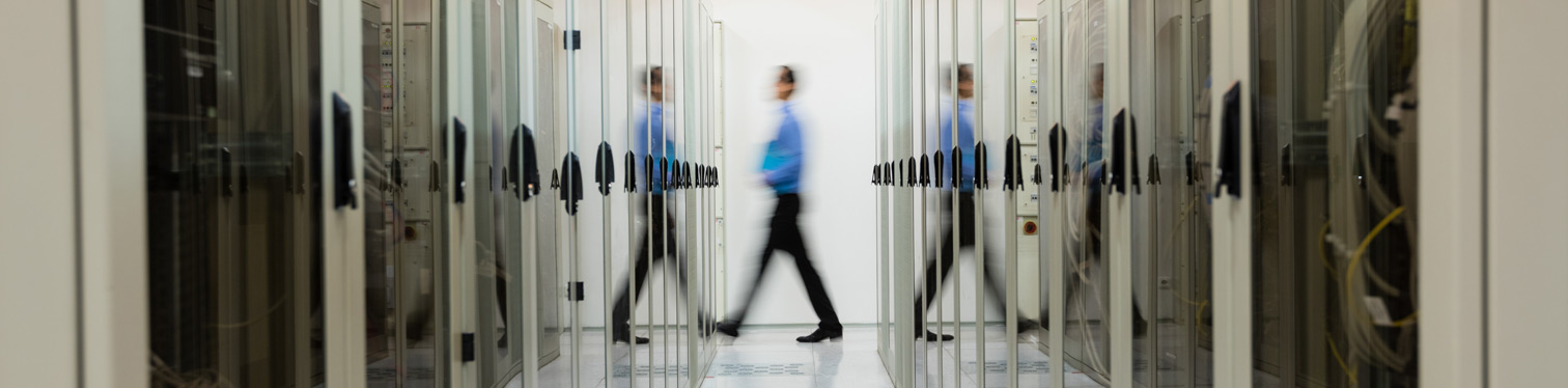Person walking through a brightly lit data center aisle lined with glass-enclosed server racks on both sides, illustrating modern IT infrastructure and enterprise technology operations.