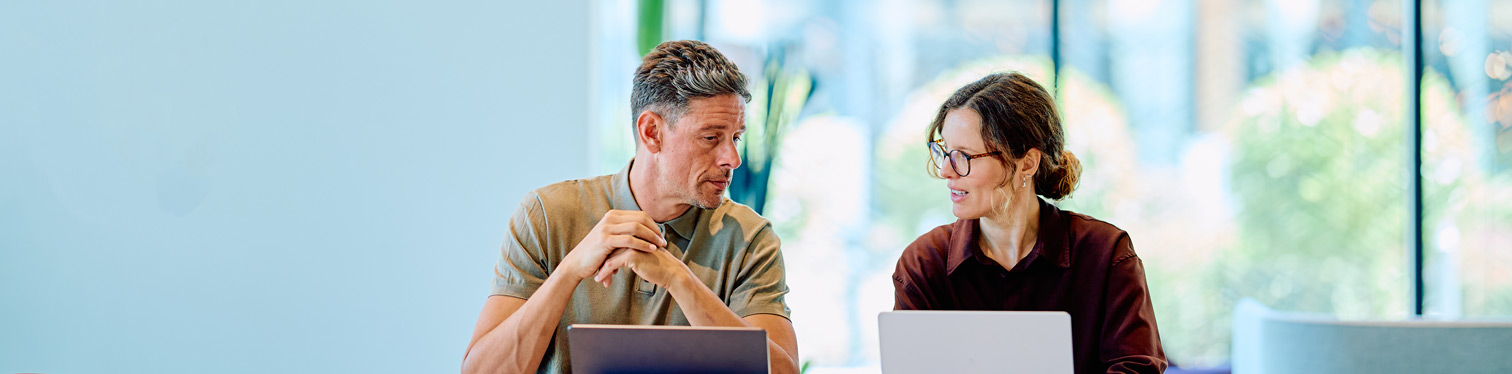 Two professionals seated at a table with laptops, discussing strategy in a modern office setting with large windows and natural light.