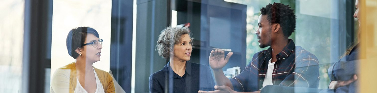 Three professionals seated in a modern office meeting space, engaged in discussion around a table with a laptop and coffee cup visible. The image is photographed through glass walls, creating reflective layers that emphasize collaboration and workplace communication.