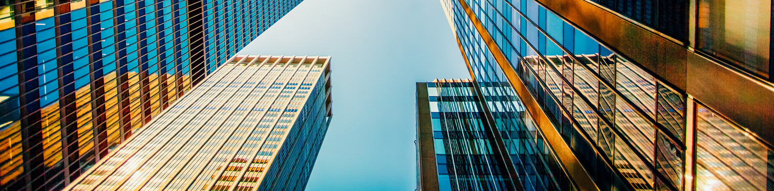 Upward view of several modern high-rise office buildings with reflective glass facades and blue-tinted windows surrounding a clear sky, creating a dramatic urban perspective.