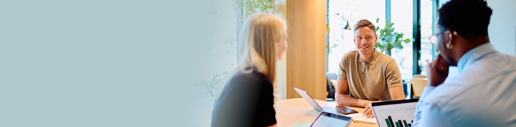 Three colleagues collaborate at a table with laptops and a tablet displaying a bar chart, discussing business data in a modern office.