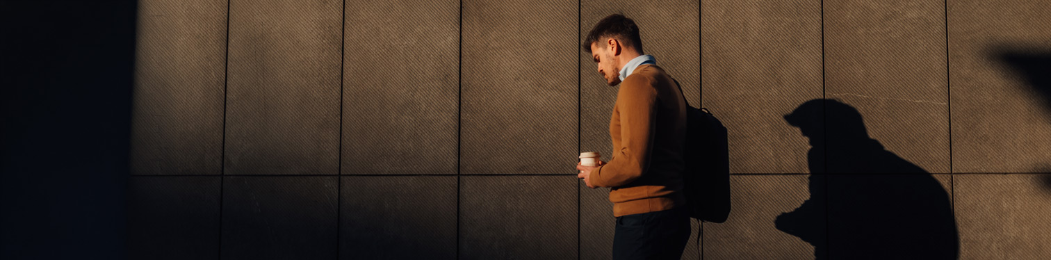 A person wearing a backpack walks alongside a large wall while holding a takeaway coffee cup. Strong sunlight casts a prominent shadow on the wall, creating a visual contrast between the person and their silhouette.
