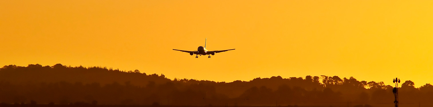 Silhouette of a commercial airplane descending for landing at sunset, with a hazy orange sky and tree-lined horizon in the background.