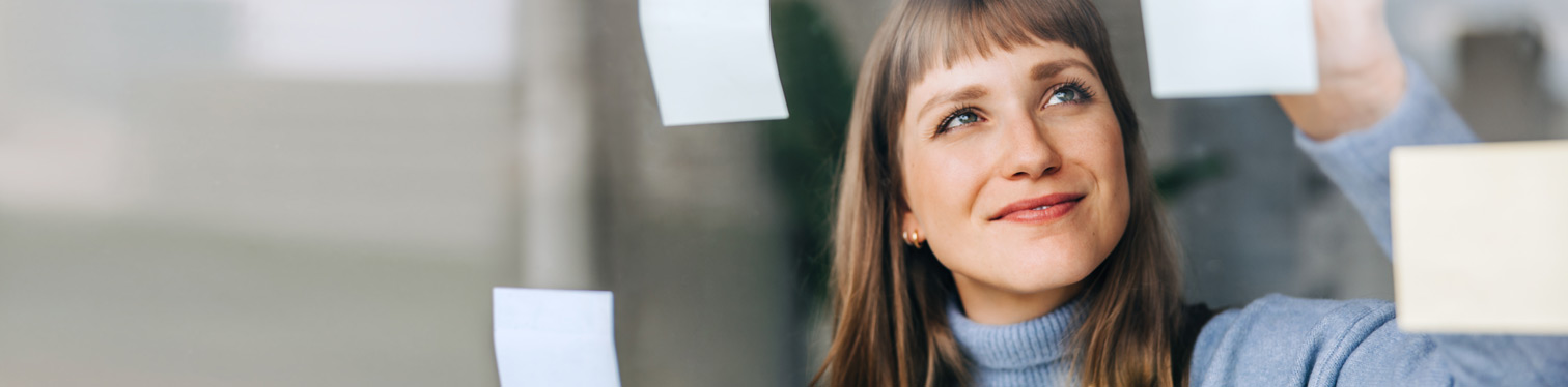 A woman in a light blue sweater places sticky notes on a glass wall while looking upward, organizing ideas or planning a project.
