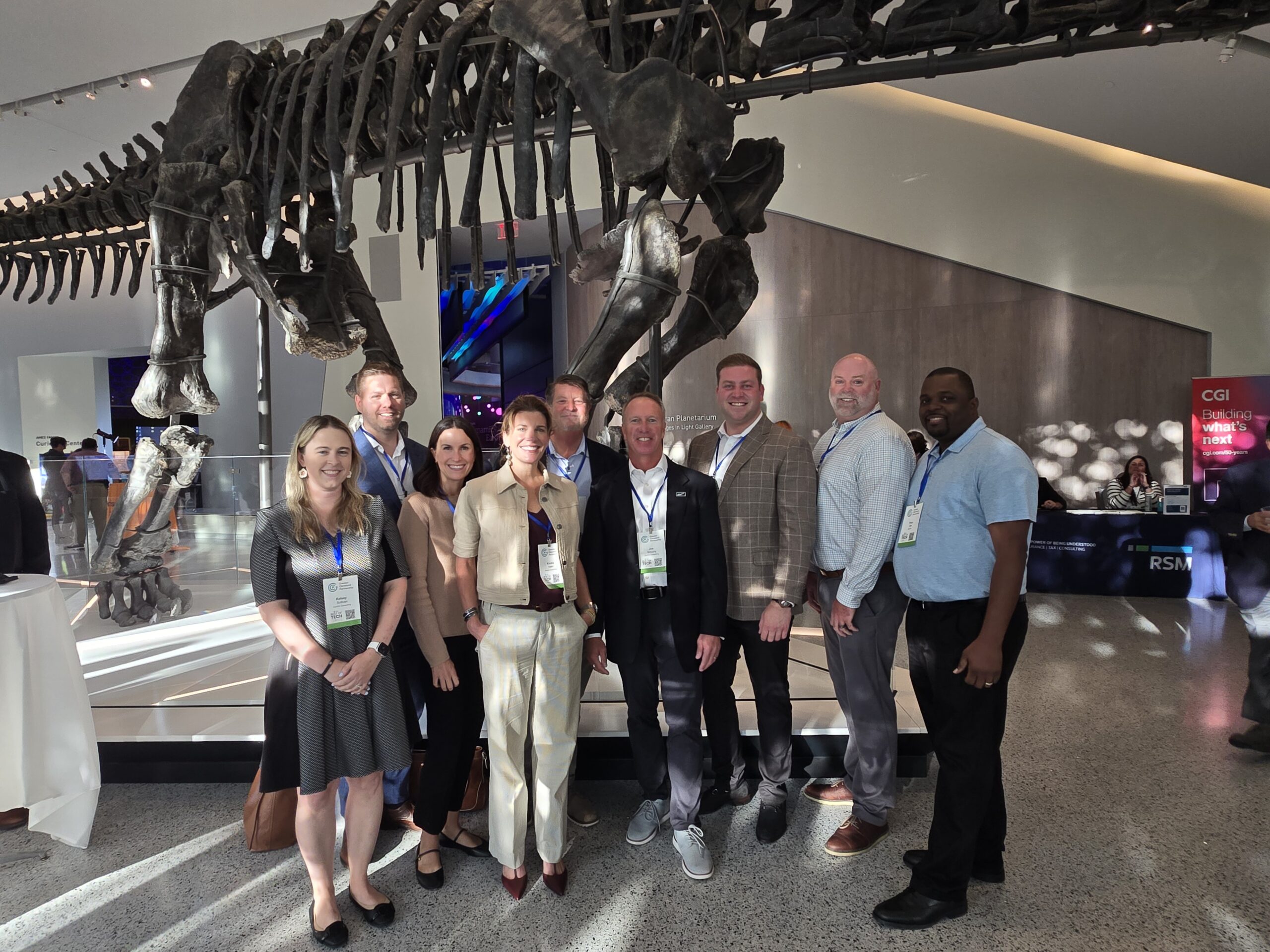 Ten Centric Consulting team members wearing event lanyards pose beneath a large dinosaur skeleton in a museum atrium during the Best of Tech event.