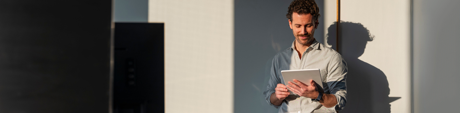 Project manager in a light button-down shirt smiles while reviewing work on a tablet, standing in warm sunlight against a modern office wall.