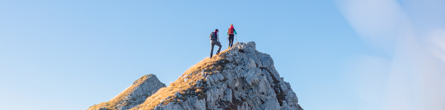 Two hikers reach the summit of a rocky mountain ridge under a clear blue sky, with golden grass lit by the sun.