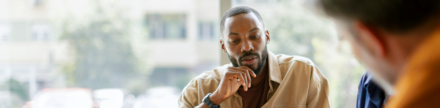 A man in a tan jacket rests his hand on his chin and looks down thoughtfully during a meeting, with a colleague blurred in the foreground.