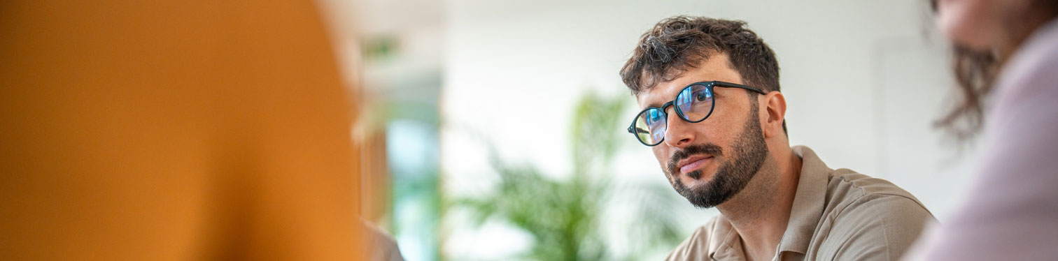 Business professional with glasses and a beard listens attentively during a meeting with colleagues, discussing ChatGPT questions for executives.