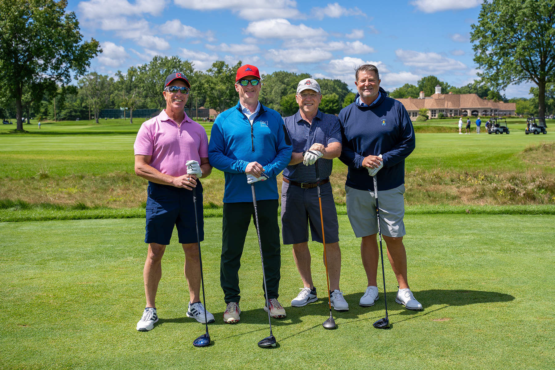 our golfers in caps and golf attire stand on a sunny tee box holding drivers, with a fairway, trees, and clubhouse behind them.