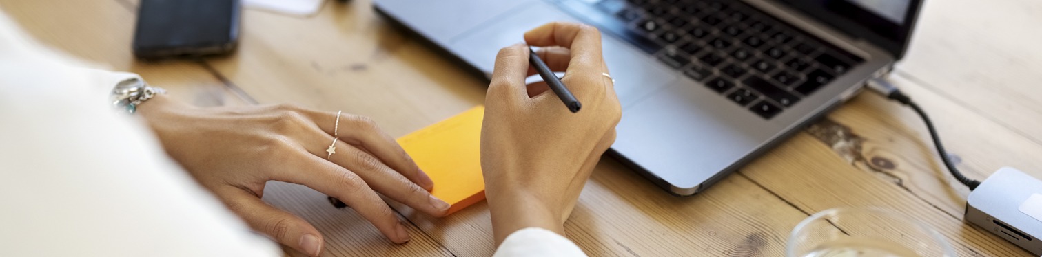 Close-up of a woman’s hands at a wooden desk, holding a stylus over an orange sticky note beside an open laptop.