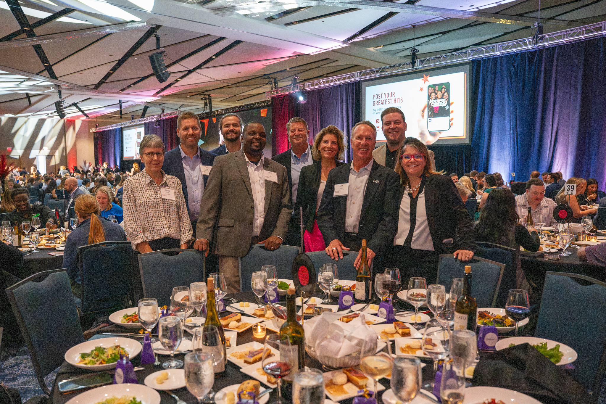 Nine Centric Consulting team members stand behind their dinner table in a large, purple-lit ballroom at the North Coast 99 awards event, with a presentation screen in the background.