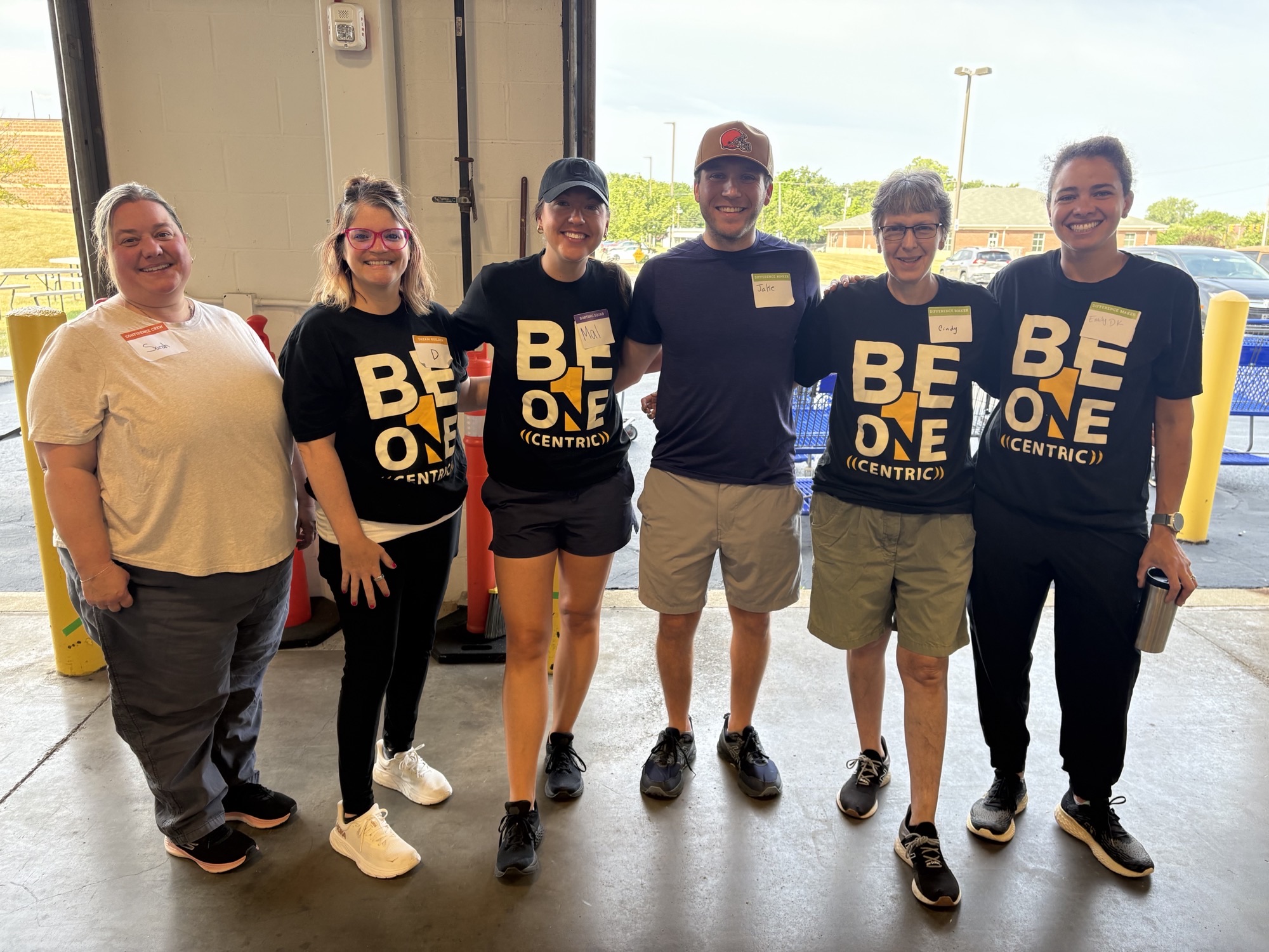 Six Centric Consulting volunteers, most wearing black “Be One Centric” T-shirts and name tags, smile together at an open warehouse loading dock.