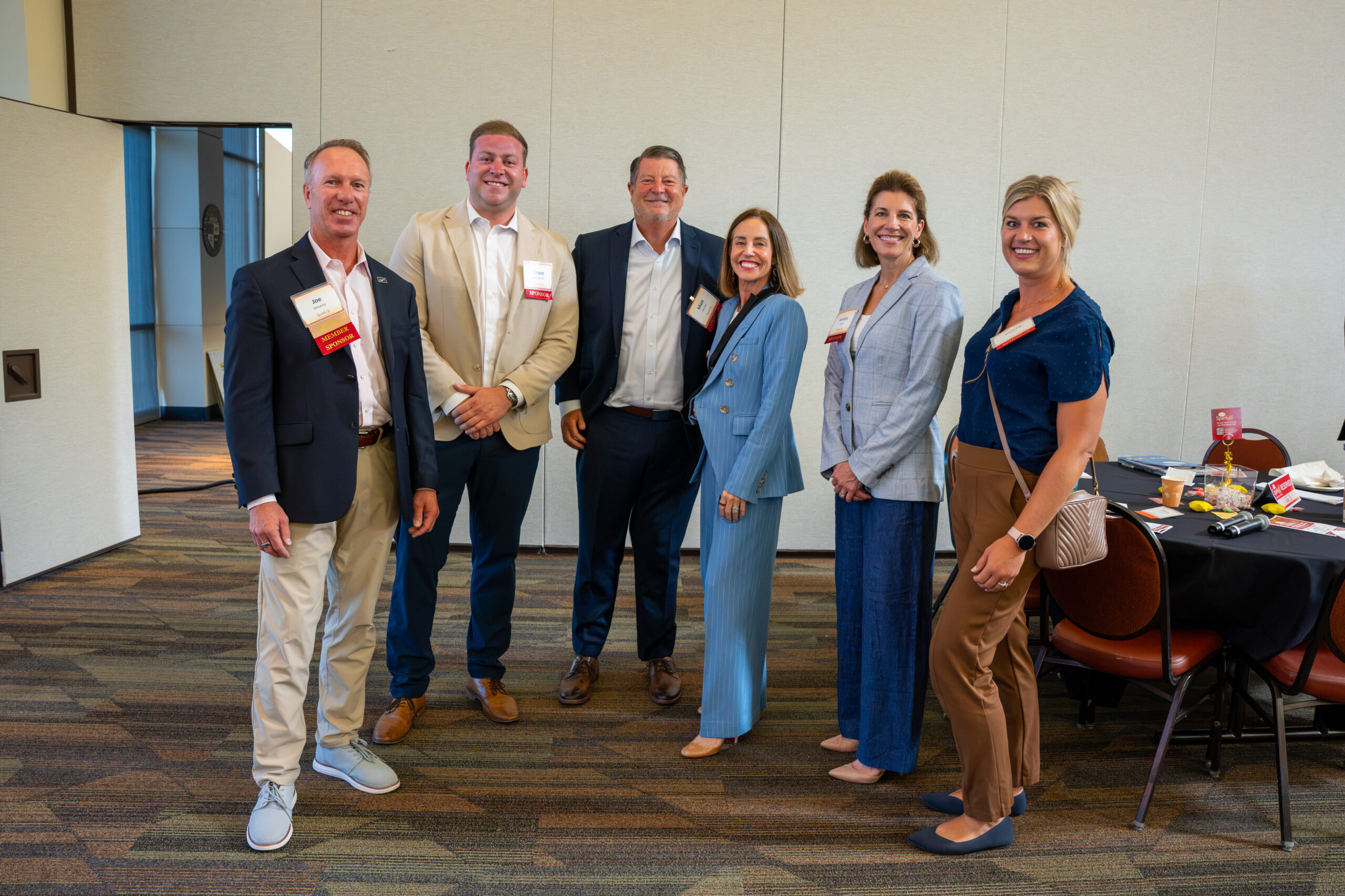Six Centric Consulting team members in business attire, several wearing member sponsor ribbons on their name badges, pose together in a conference room beside a dining table.